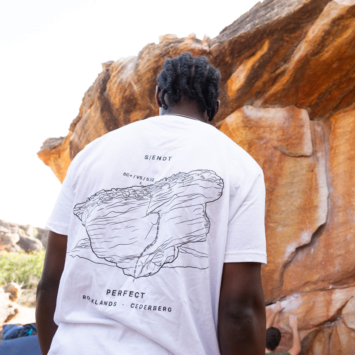 A person viewed from behind wearing a white t-shirt with a black line-art boulder graphic and text on the back, standing outdoors in front of a large orange-brown rock formation in Cederberg South Africa
