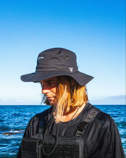 Person wearing a black bucket hat and black outfit standing by the ocean with clear blue sky.
